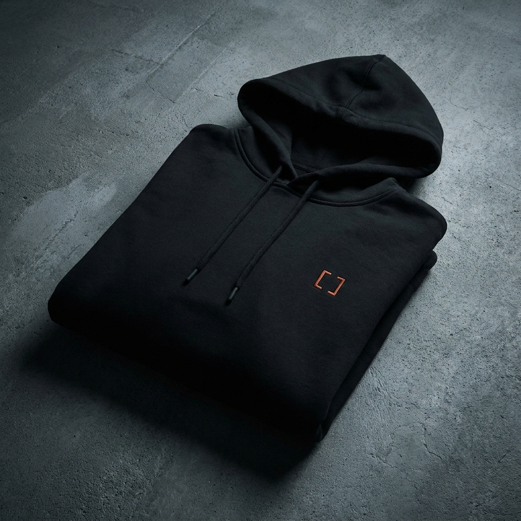 Black pullover hoodie with orange bracket emblem, folded on dark concrete