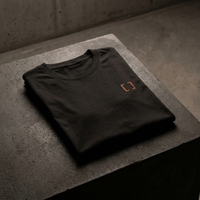 Black crew t-shirt with orange bracket emblem, folded on dark concrete