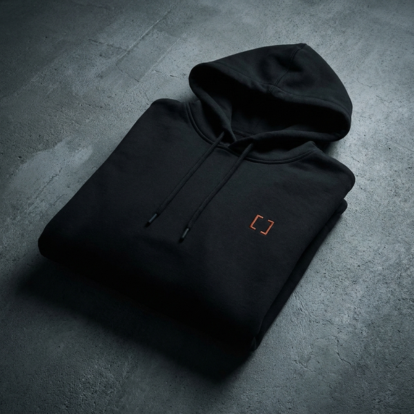 Black pullover hoodie with orange bracket emblem, folded on dark concrete